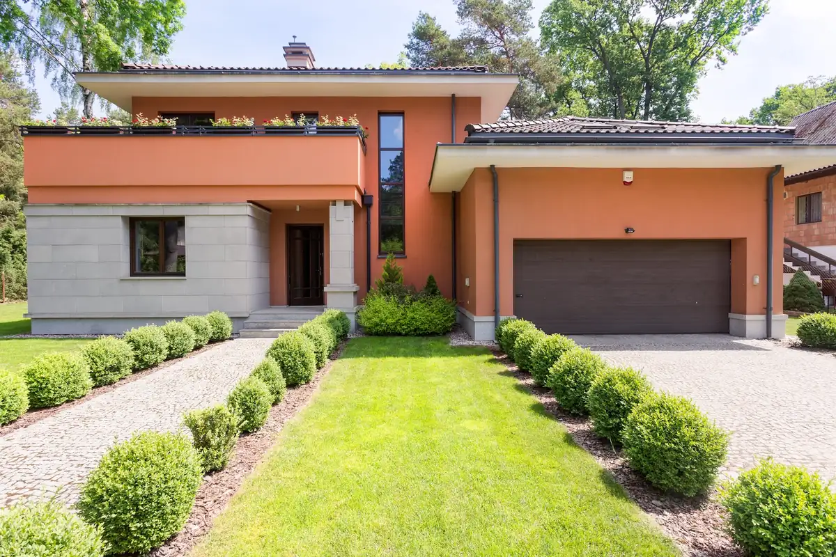 Modern two story home painted bright orange with a gray stone base, dark garage door and a green lawn lined with round shrubs