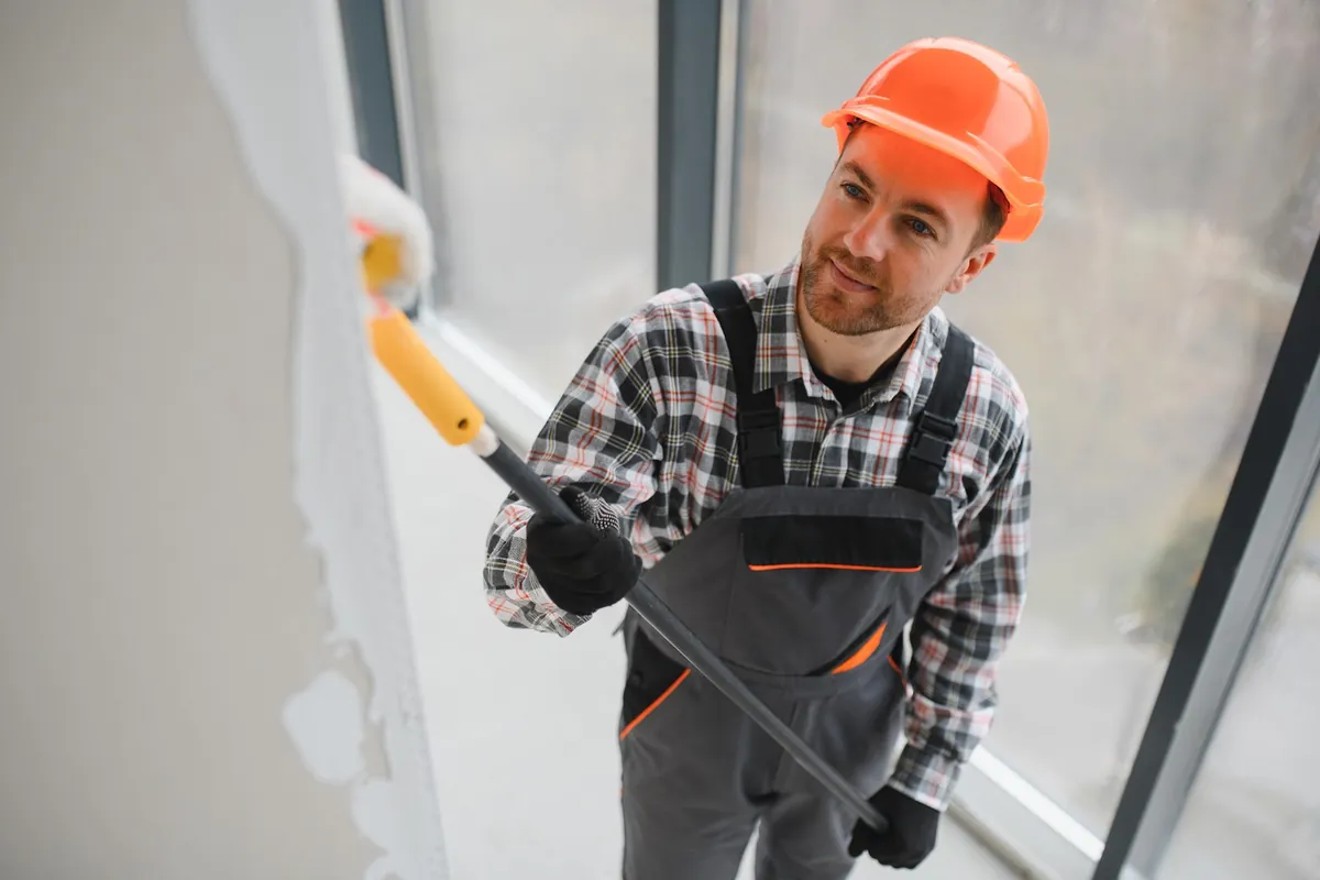 Painter in a hard hat rolling paint on an office wall beside windows
