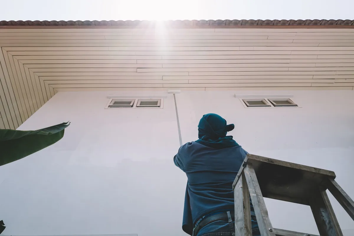 Painter on a ladder rolling white paint onto a house wall