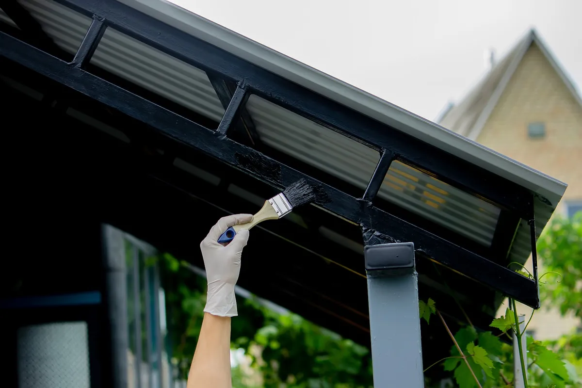 Gloved hand brushing black paint onto a metal awning frame