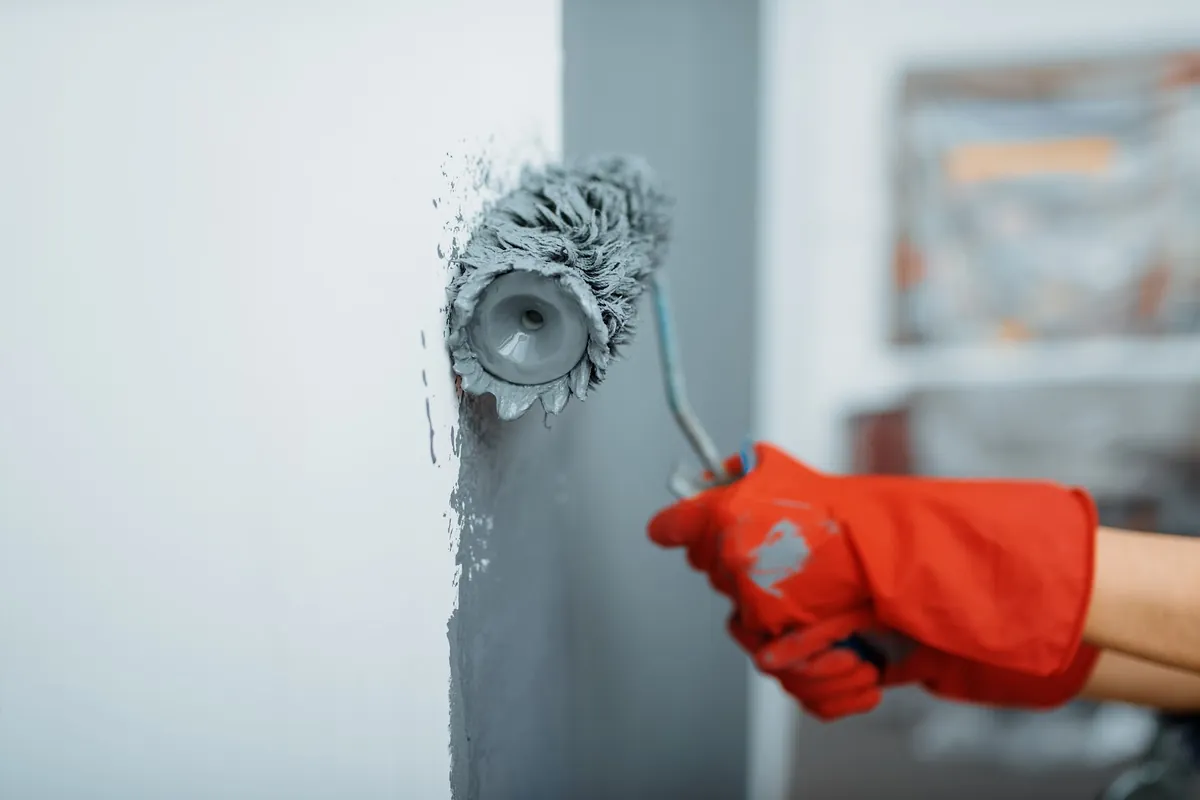 Gloved hand rolling gray paint into a wall corner